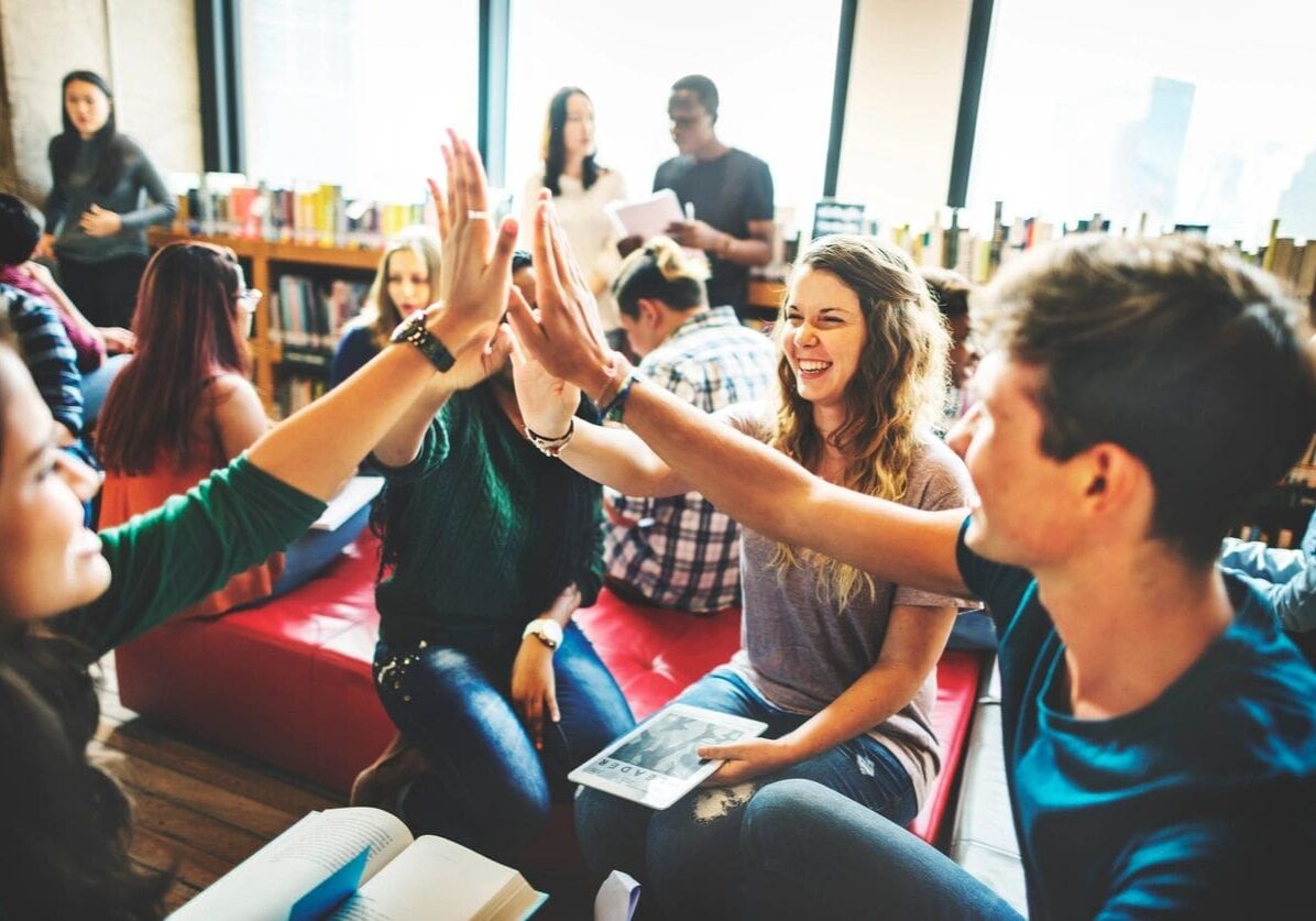 fm=f_pYEqm8V A group of diverse young people giving high fives and smiling in a casual indoor setting.